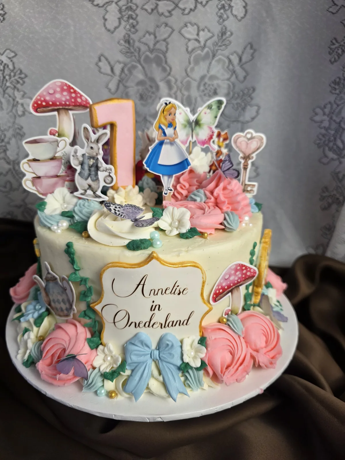 White buttercream cake decorated with Alice character, white rabbit, mushrooms, butterfly, key, teacups, pink number one, pastel flowers, and a blue fondant bow.