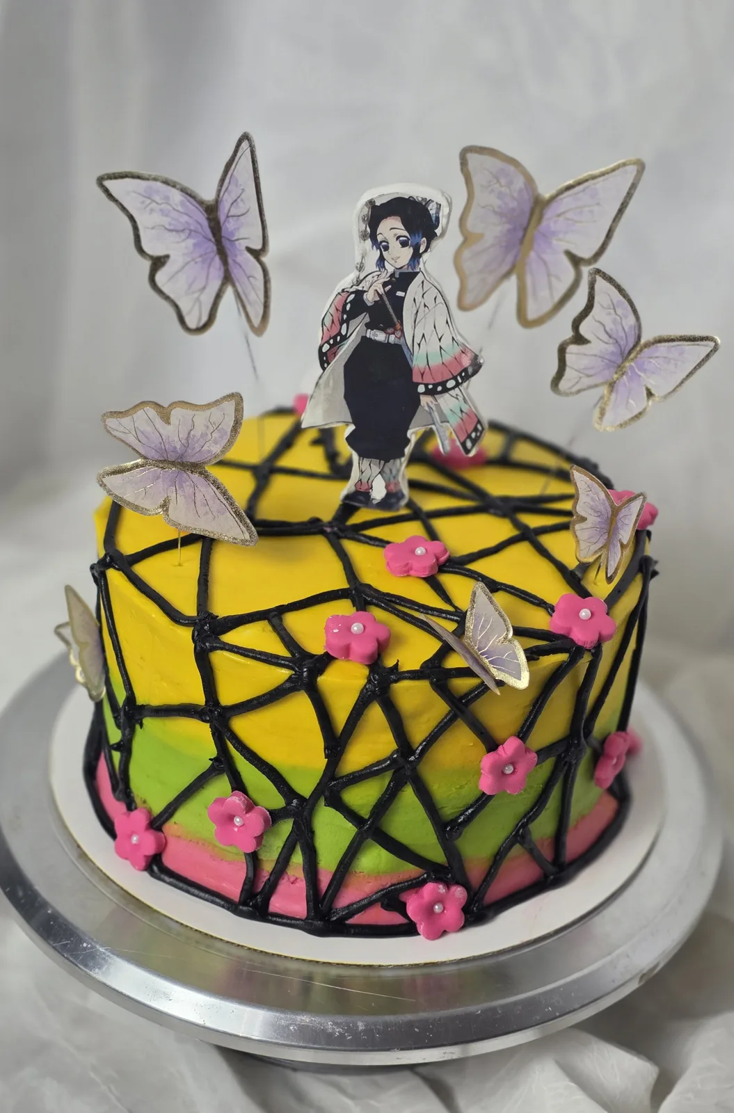 Round cake with yellow, green, and pink fondant layers, black piped geometric web pattern, pink flowers, and anime figure topper with purple butterfly accents.