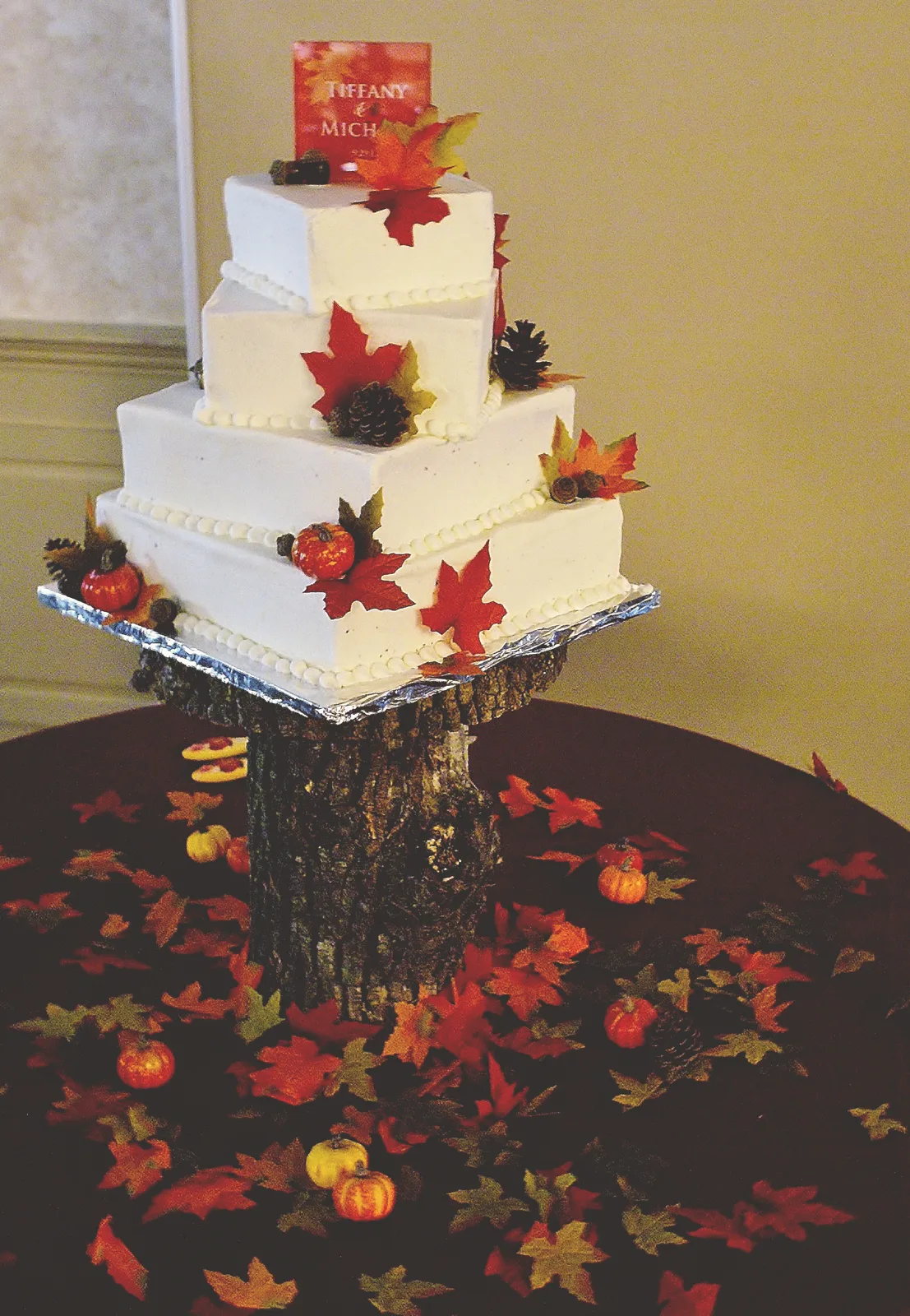 Four tier white square wedding cake adorned with red and orange fall leaves, pine cones, and mini pumpkins, displayed on a rustic tree stump base.
