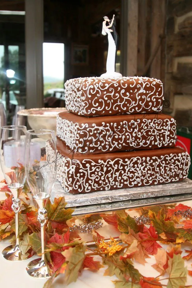 Three tier square chocolate cake with intricate white piped scrollwork on each tier, topped with white bride and groom figurine on silver base surrounded by autumn leaves.