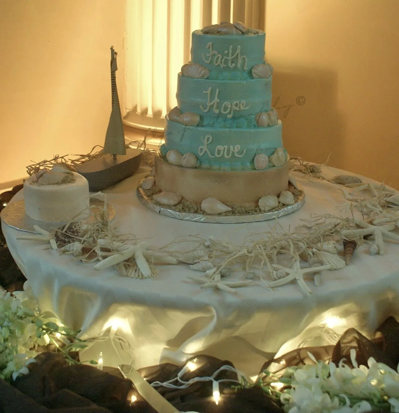 Three tiered turquoise and sand colored cake inscribed with Faith, Hope, and Love adorned with seashells on a table scattered with shells and starfish.