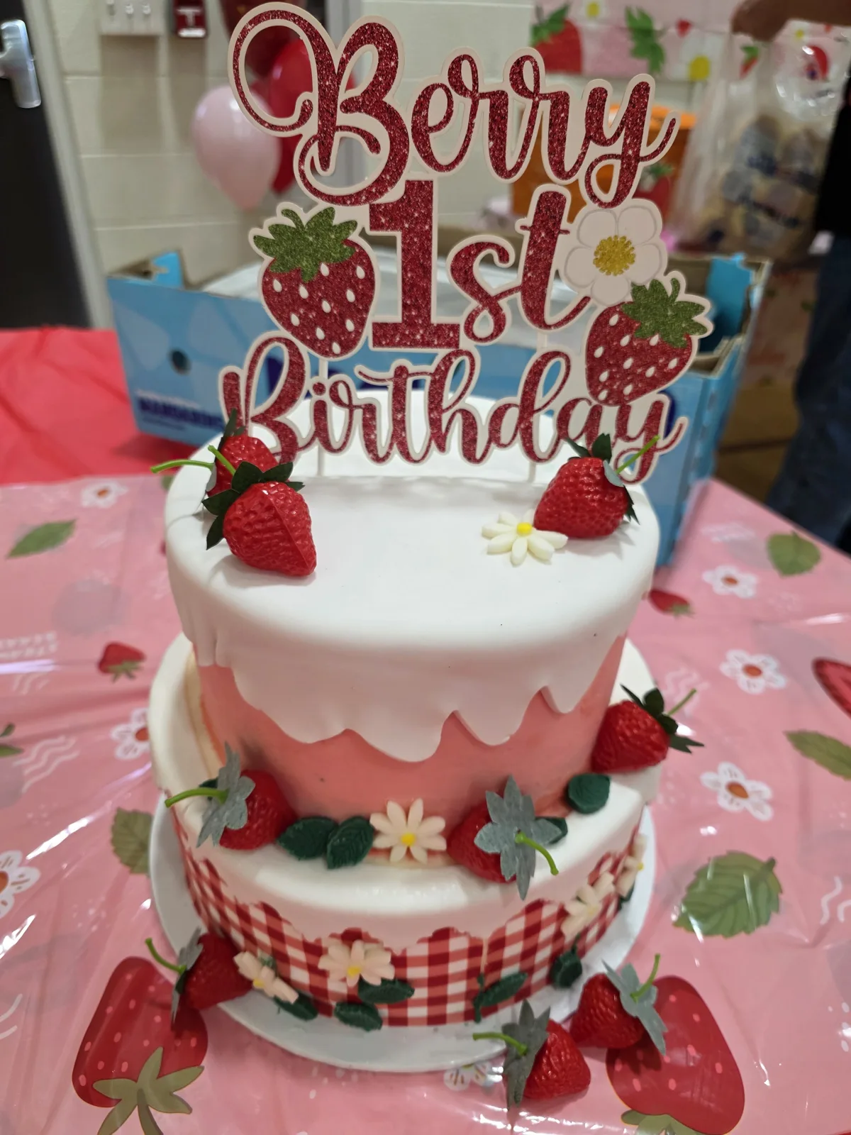 Two tier white and pink drip cake decorated with fresh strawberries, small white daisy flowers, and a red gingham ribbon, topped with a red glitter "Berry 1st Birthday" sign.