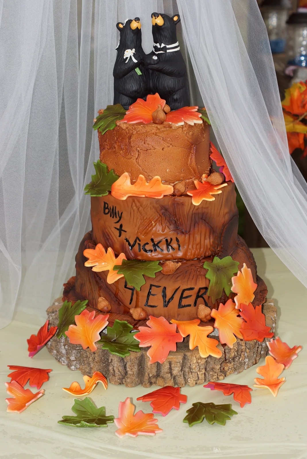 Three tiered chocolate frosted wedding cake with wood bark texture, topped with two black bear figurines and decorated with orange, red, and green fondant autumn leaves.