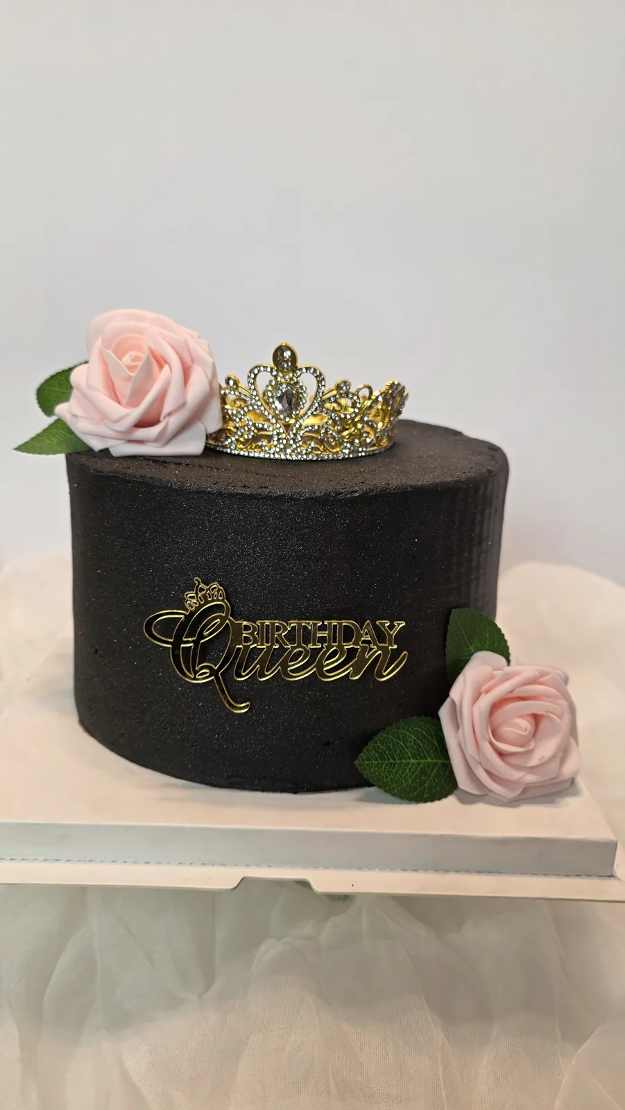 Tall round dark chocolate frosted cake topped with a rhinestone tiara, pink roses with green leaves, and a gold Birthday Queen topper.