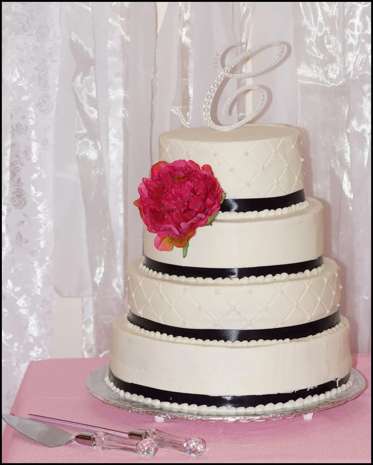 Four tier ivory fondant wedding cake decorated with black satin ribbon trim, quilted lattice pattern, pearl beading, a large magenta peony, and a rhinestone letter topper.