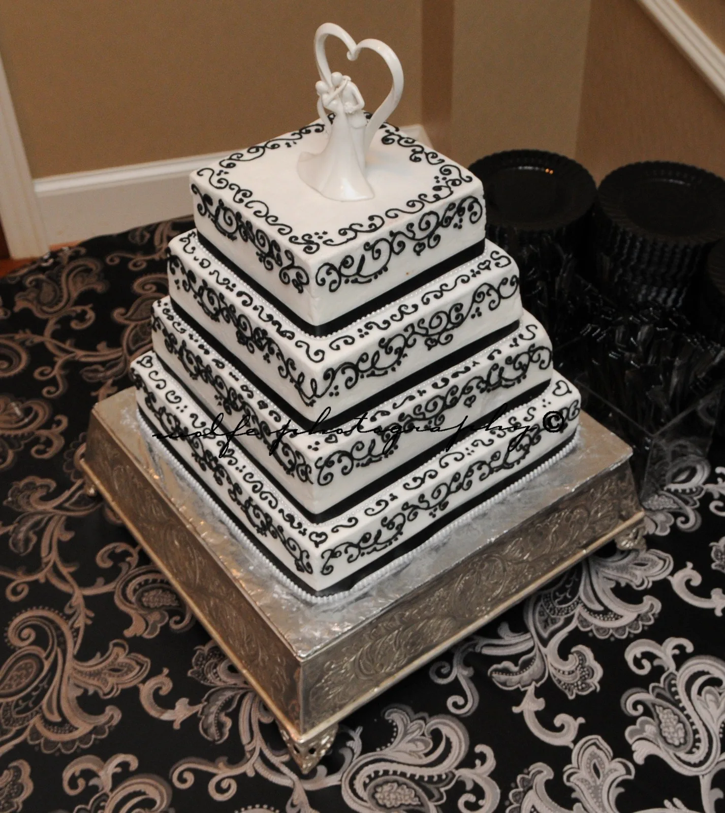 Four tiered square white wedding cake decorated with intricate black scrollwork piping, topped with a bride and groom figurine beneath a heart shaped arch.