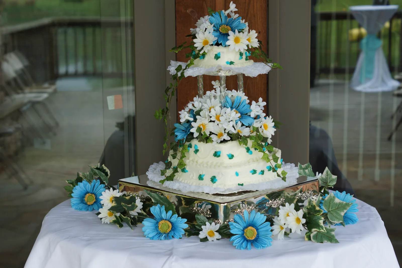 Two tier white wedding cake adorned with blue and white daisies, green ivy, and scattered blue floral accents on a mirrored base.