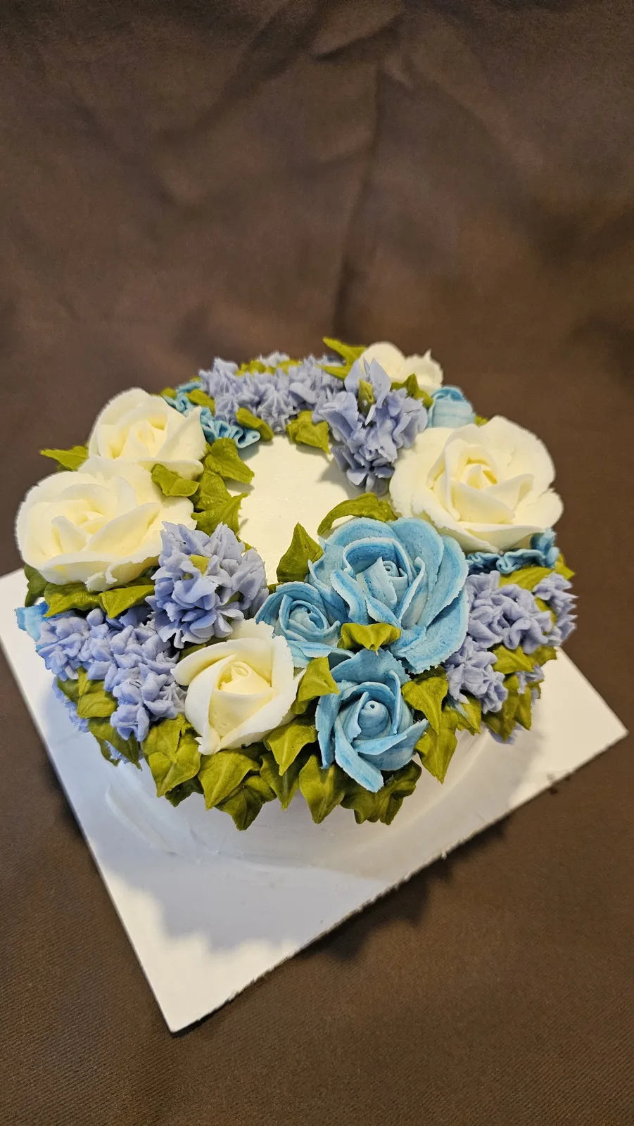 Round cake topped with piped buttercream roses and hydrangeas in white, blue, and lavender hues surrounded by green leaves.