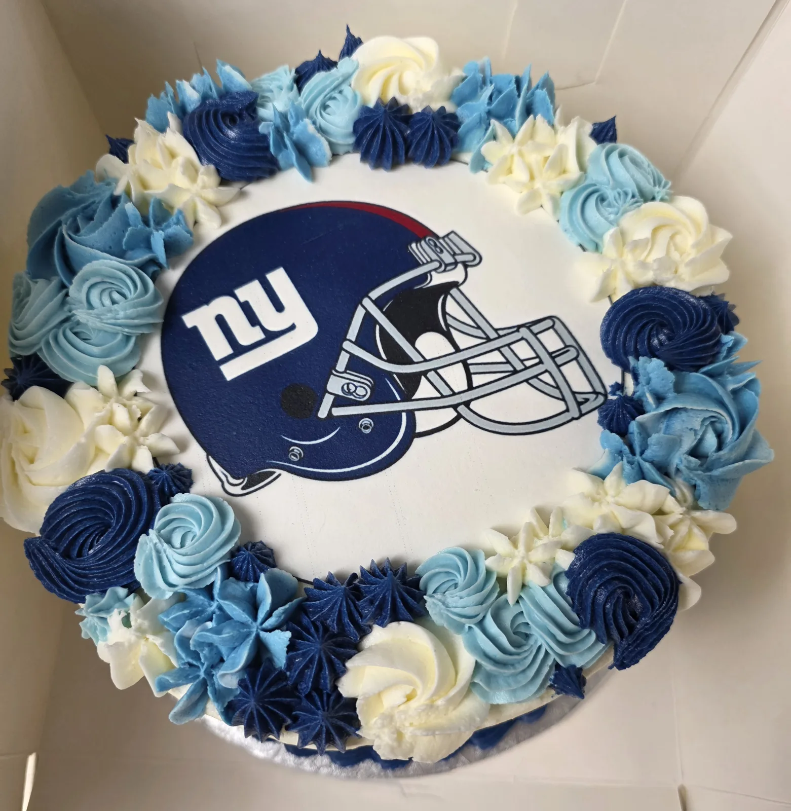 Round white frosted cake featuring a blue football helmet decal, bordered with piped rosettes in navy, light blue, and white buttercream.