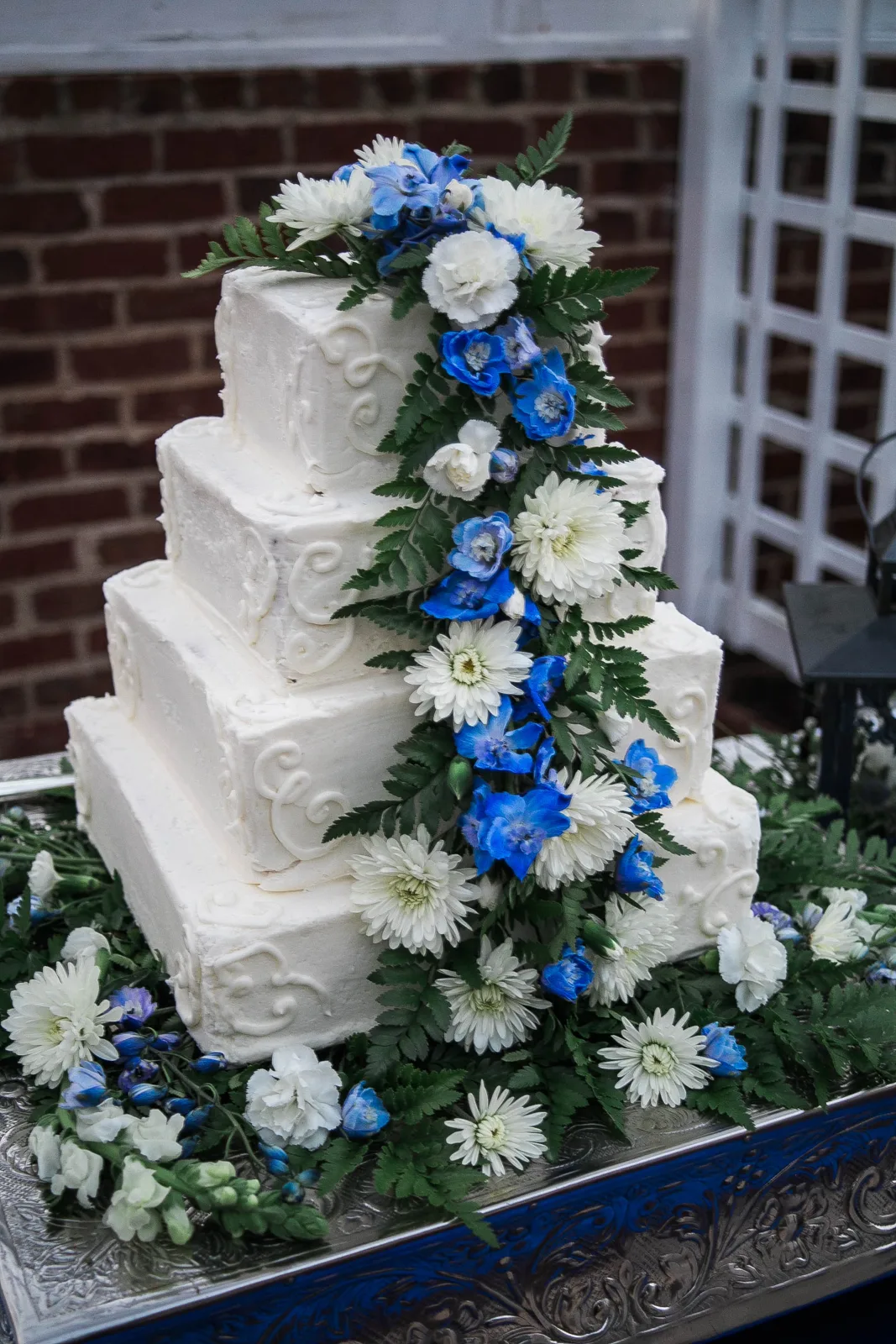 Four tiered square white wedding cake with piped scrollwork and a cascading arrangement of blue delphiniums, white chrysanthemums, and green ferns.