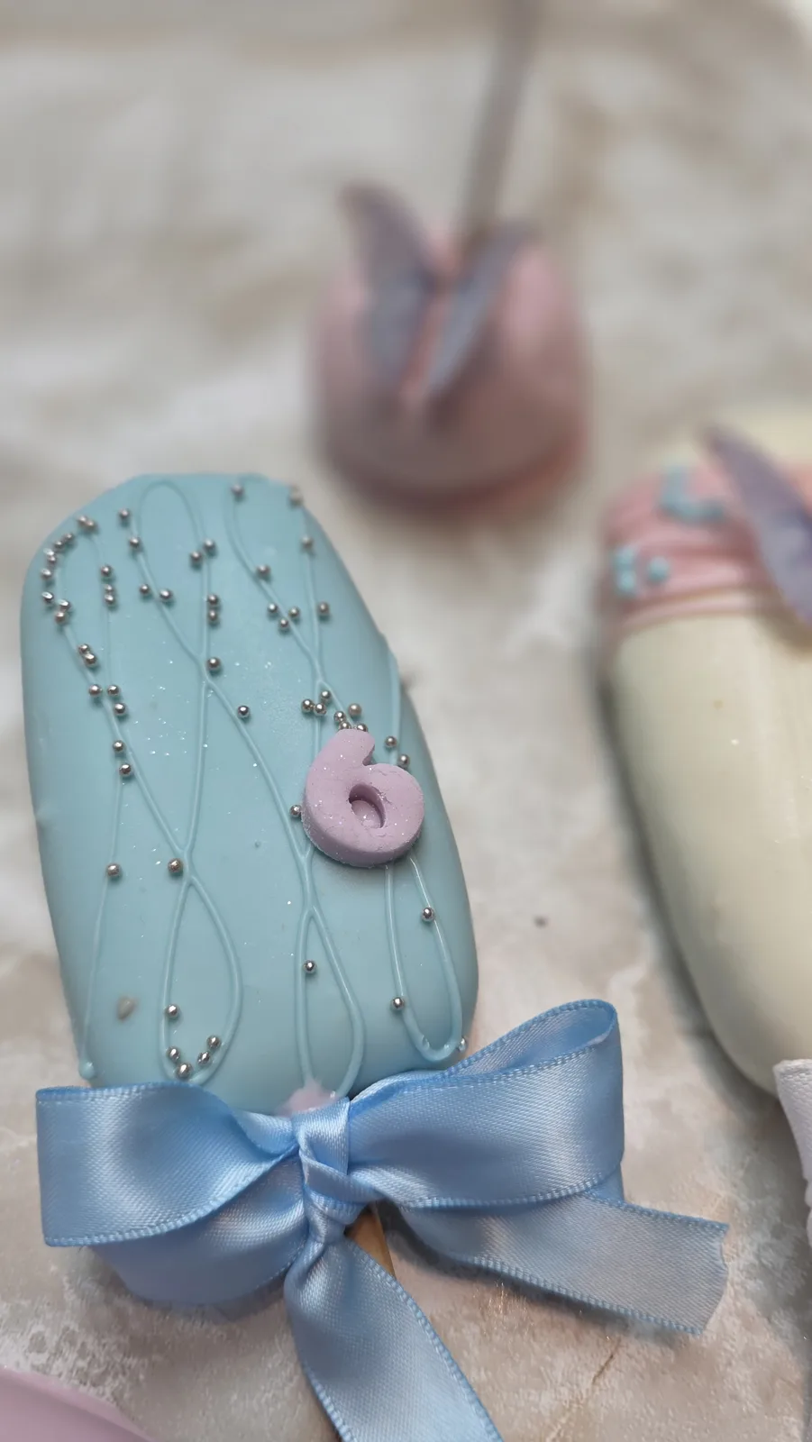 A light blue chocolate coated cakesicle with white chocolate drizzle, silver pearl sprinkles, a lavender number six, and a blue satin bow tied at the stick.