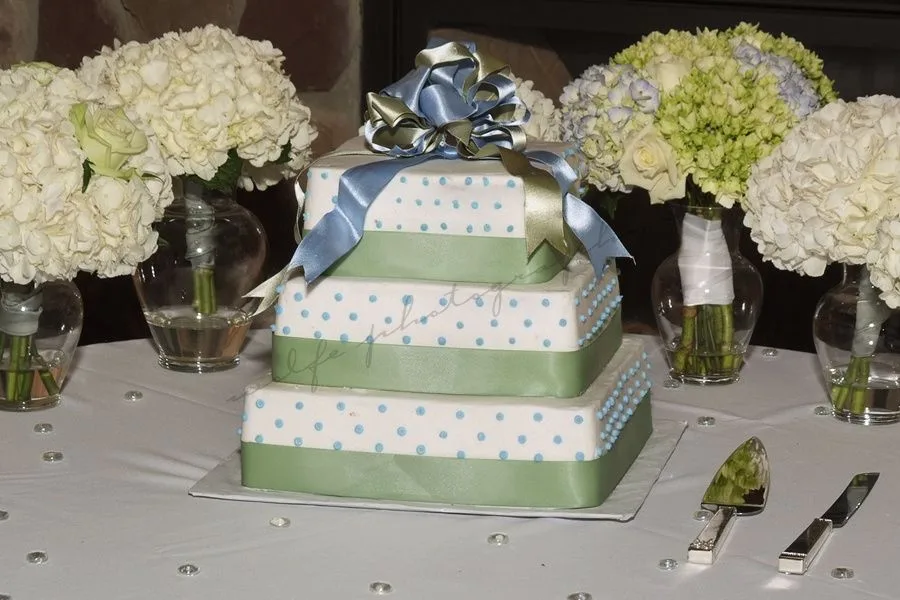 Three tiered square white fondant cake decorated with light blue polka dots, sage green ribbon borders, and topped with a layered blue and green ribbon bow.