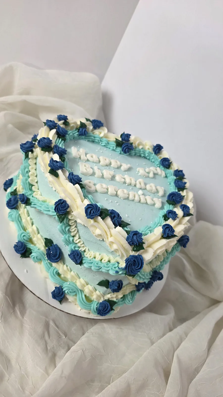 Heart shaped cake with light blue frosting, white piped ruffles, navy blue roses with green leaves, and white piped lettering on top.