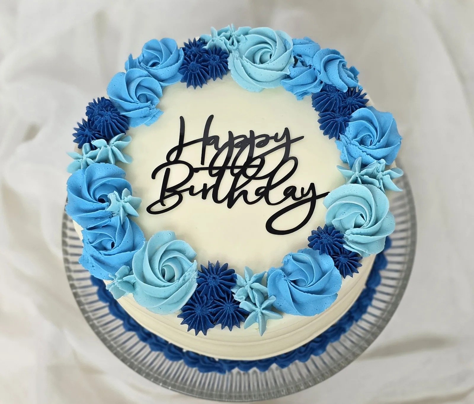 White frosted cake topped with a ring of blue and light blue buttercream roses and star shaped accents, featuring a black Happy Birthday cake topper.
