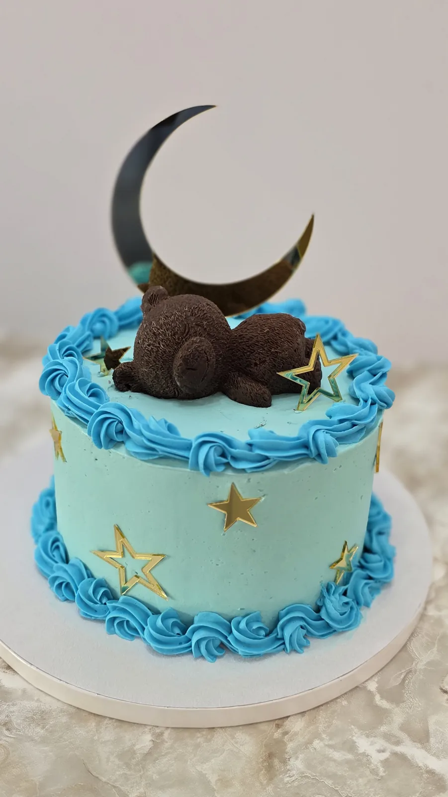 Light blue frosted round cake decorated with blue buttercream rosettes, gold star accents, a chocolate teddy bear, and a gold crescent moon topper.