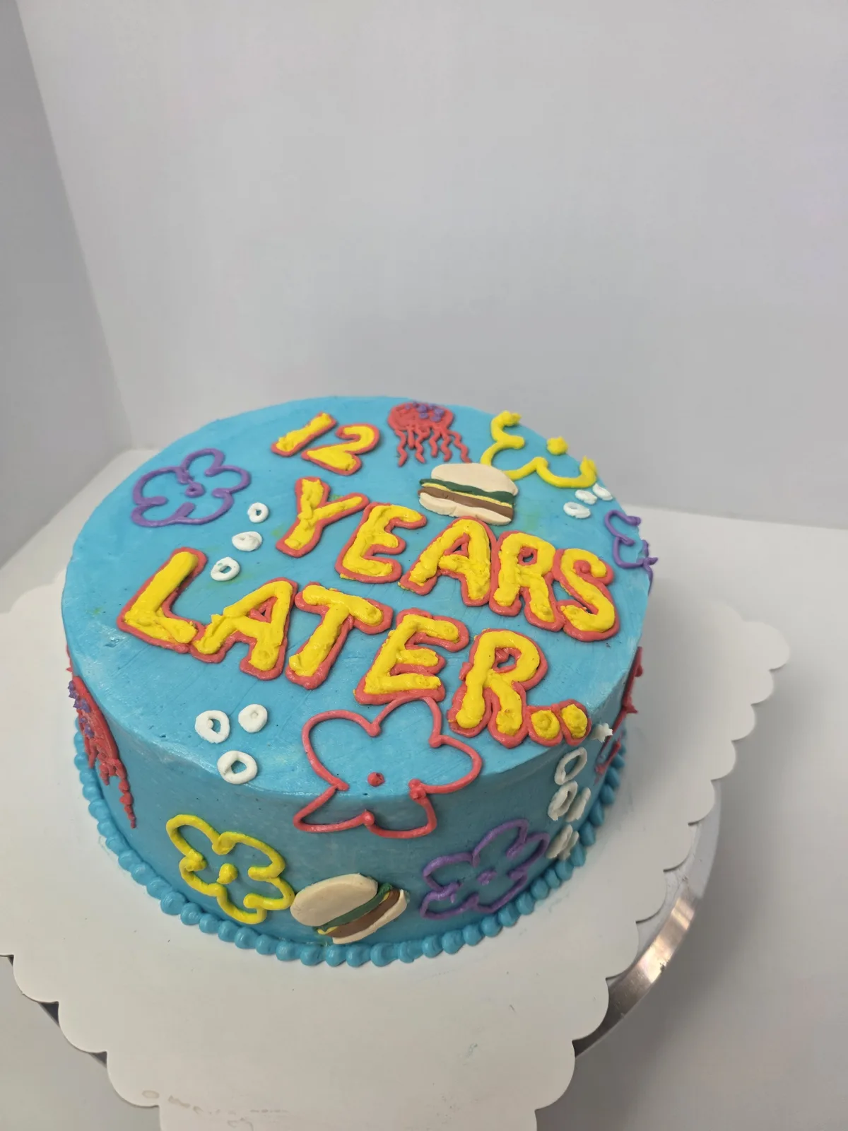 Bright blue frosted cake featuring yellow and red piped text reading twelve years later, decorated with cartoon clouds, a jellyfish, and small burger accents on a white scalloped board.