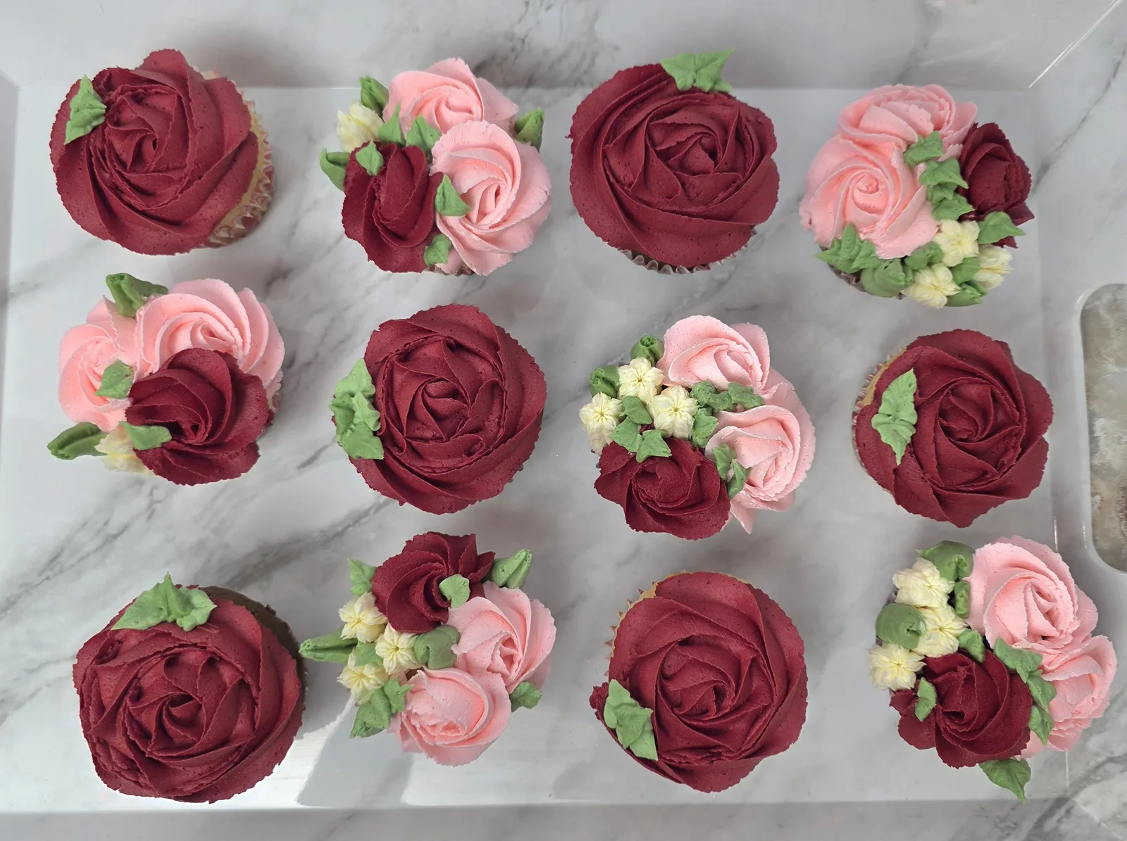 A dozen cupcakes topped with piped buttercream roses in deep red, soft pink, and cream hues with green leaves arranged on a white marble surface.