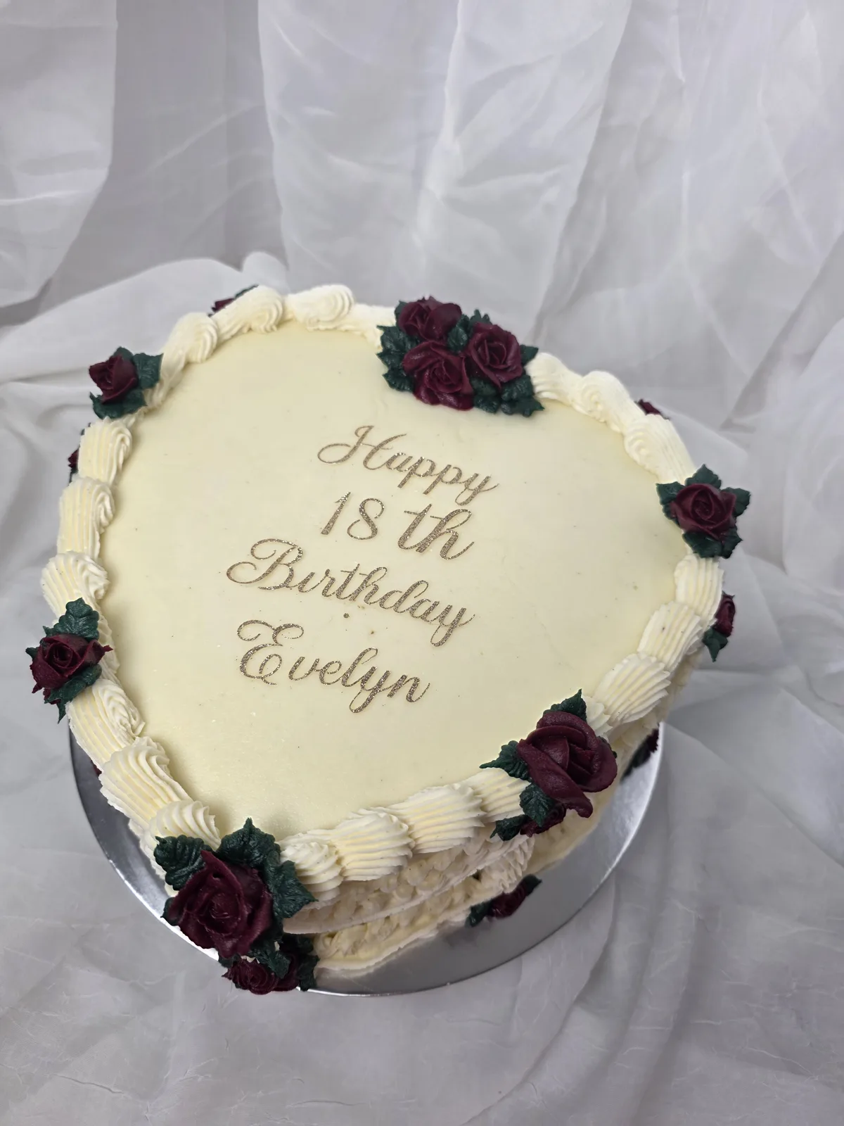 White frosted cake decorated with piped cream borders and clusters of deep burgundy roses with green leaves around the edges.