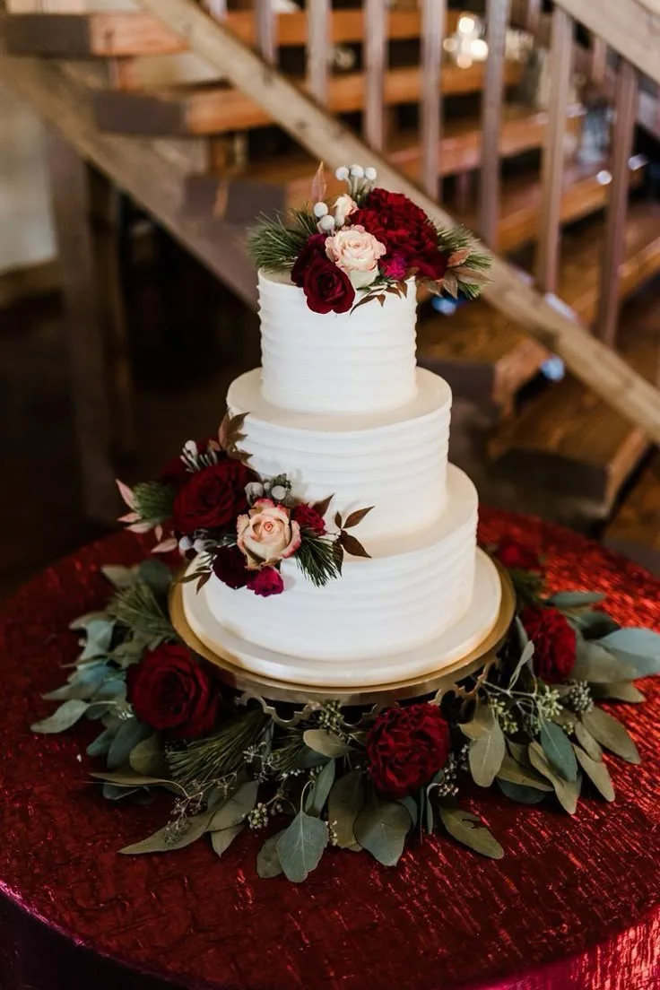 Three tier white buttercream cake with textured ridges, adorned with burgundy and blush roses, white berries, and pine, displayed on a red sequined tablecloth.