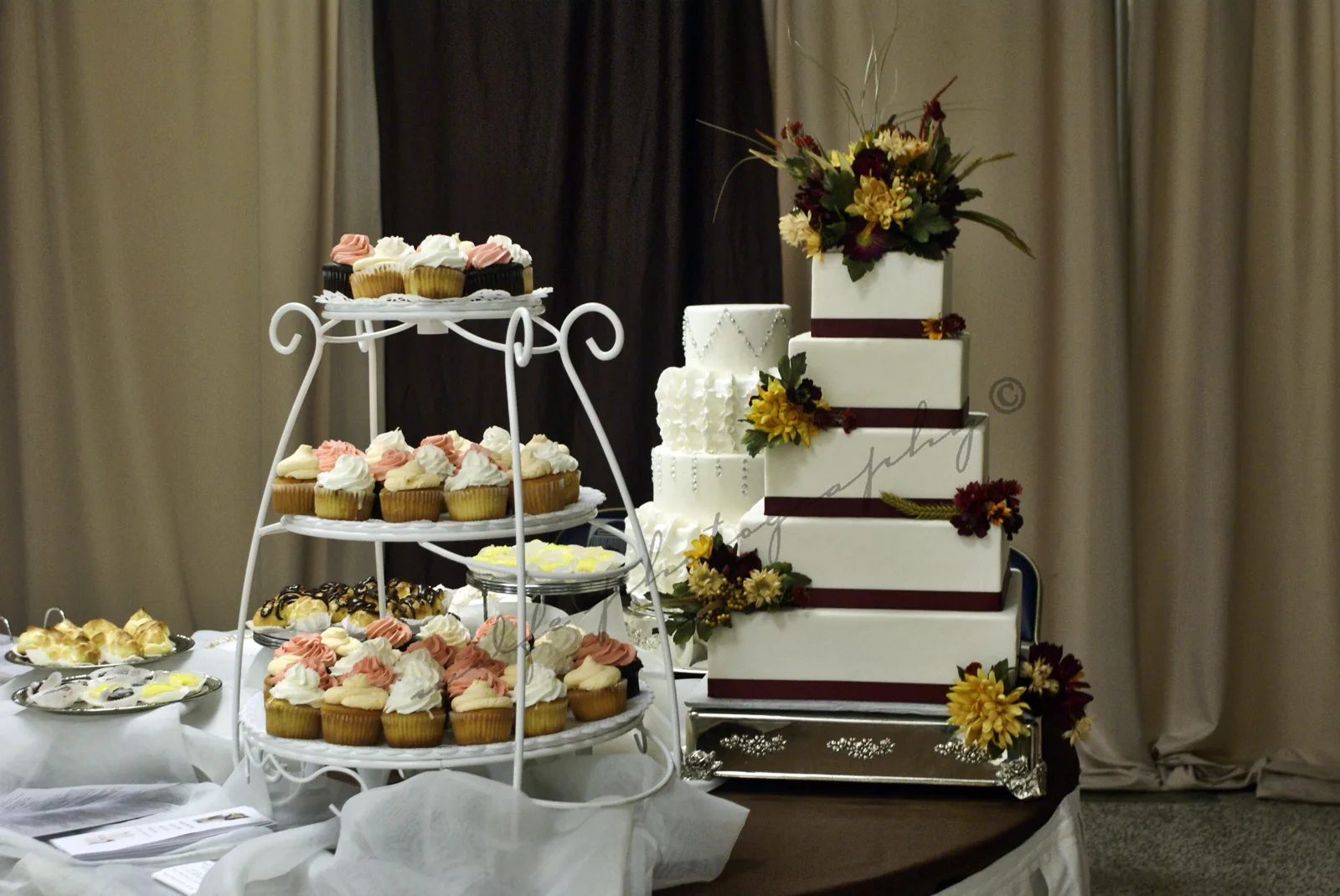 Five tiered square white wedding cake with burgundy ribbon trim, textured fondant details, and yellow sunflower and burgundy dahlia floral accents beside a tiered cupcake stand.