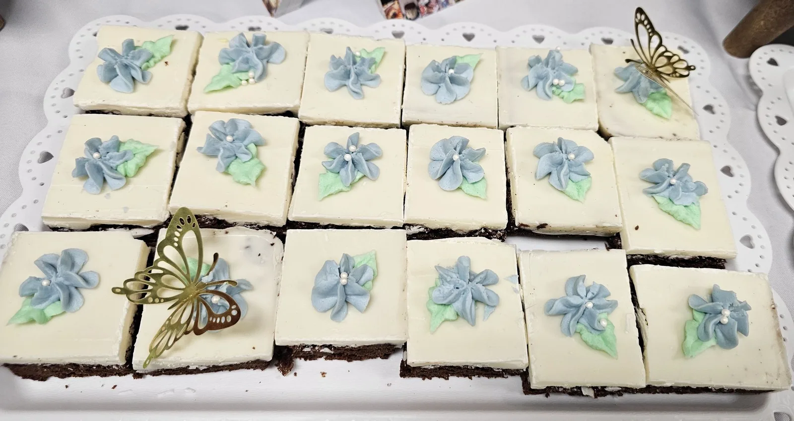 Grid of square brownie bites topped with white icing, piped blue buttercream flowers and gold butterfly picks on a scalloped tray.