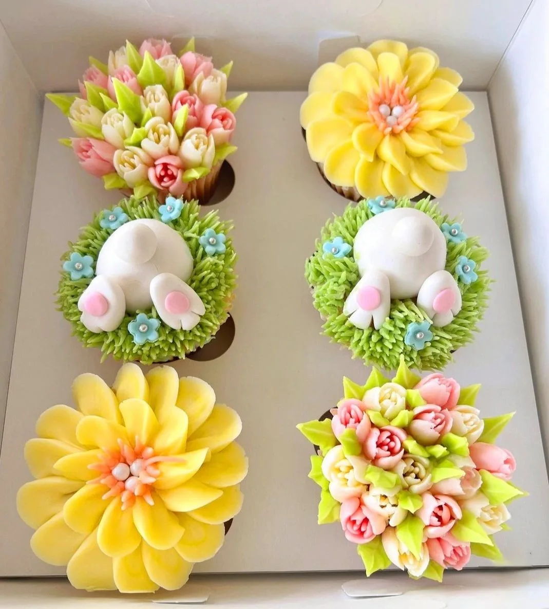 A box of six decorated cupcakes featuring yellow daisy designs, pink and cream tulip bouquets, and bunny bottoms with pink feet in green frosting.