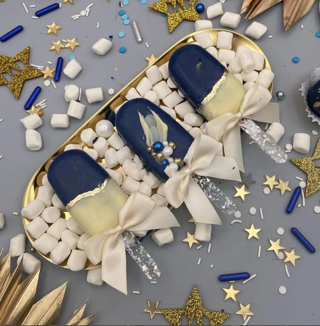 Three navy and white chocolate-dipped treats with cream bows on a gold tray surrounded by marshmallows and gold stars.