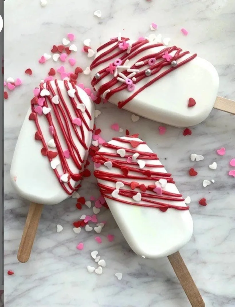 Three white chocolate-covered cakesicles on wooden sticks, drizzled with red icing and topped with pink and red sprinkles.