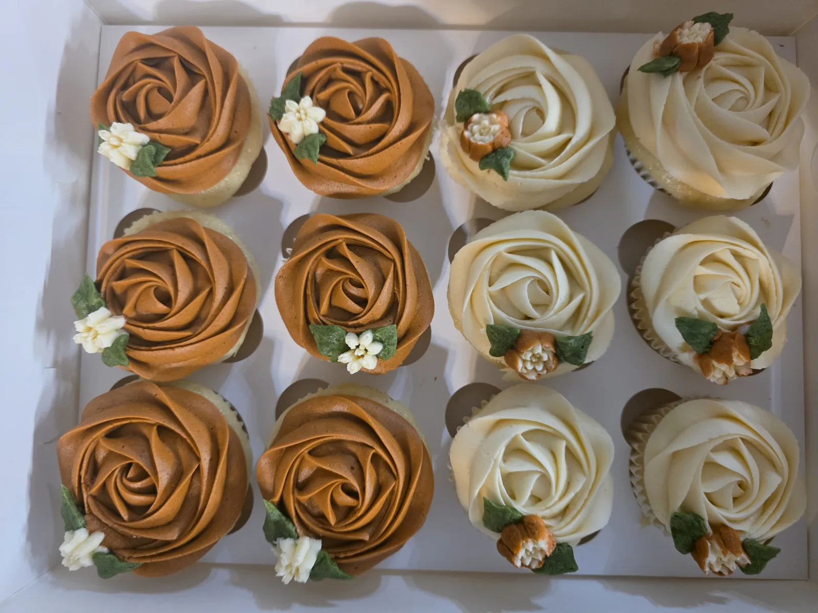 A dozen cupcakes piped as caramel and cream buttercream roses with small piped blossoms and green leaves, in a white bakery box.