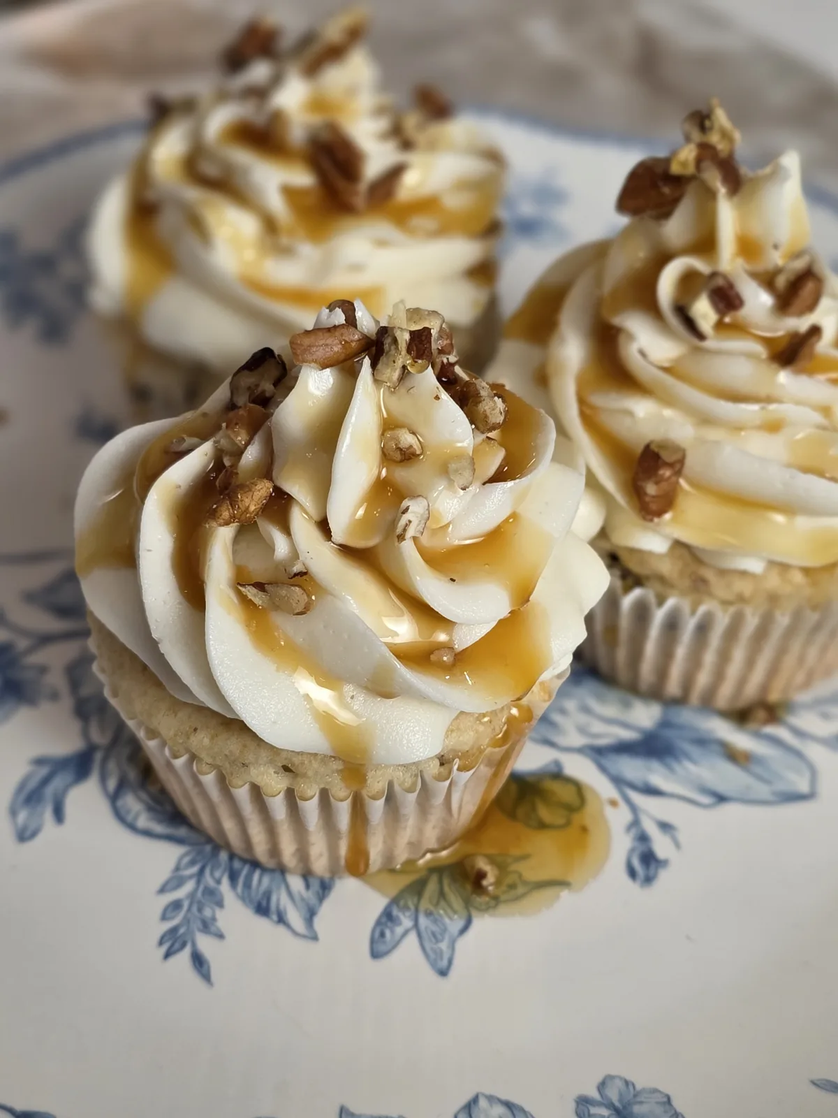 Three cupcakes with tall swirls of white frosting drizzled with golden caramel syrup and topped with chopped pecans on a blue floral plate.
