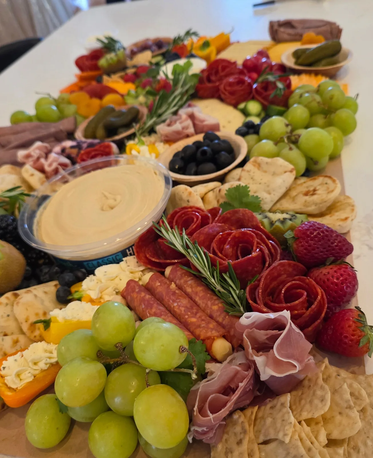 Charcuterie board featuring salami roses, prosciutto, grapes, strawberries, blueberries, kiwi, mini flatbreads, and a hummus container.