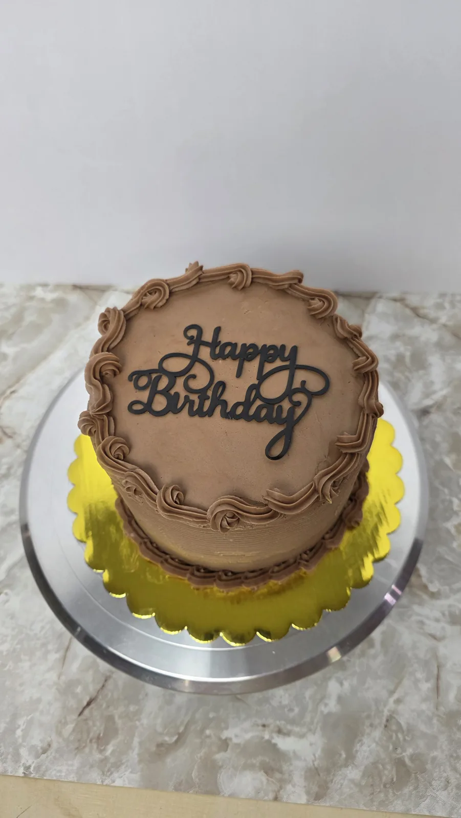 Round chocolate frosted cake with piped rosette borders and a black cursive Happy Birthday topper, displayed on a gold scalloped board and silver stand.
