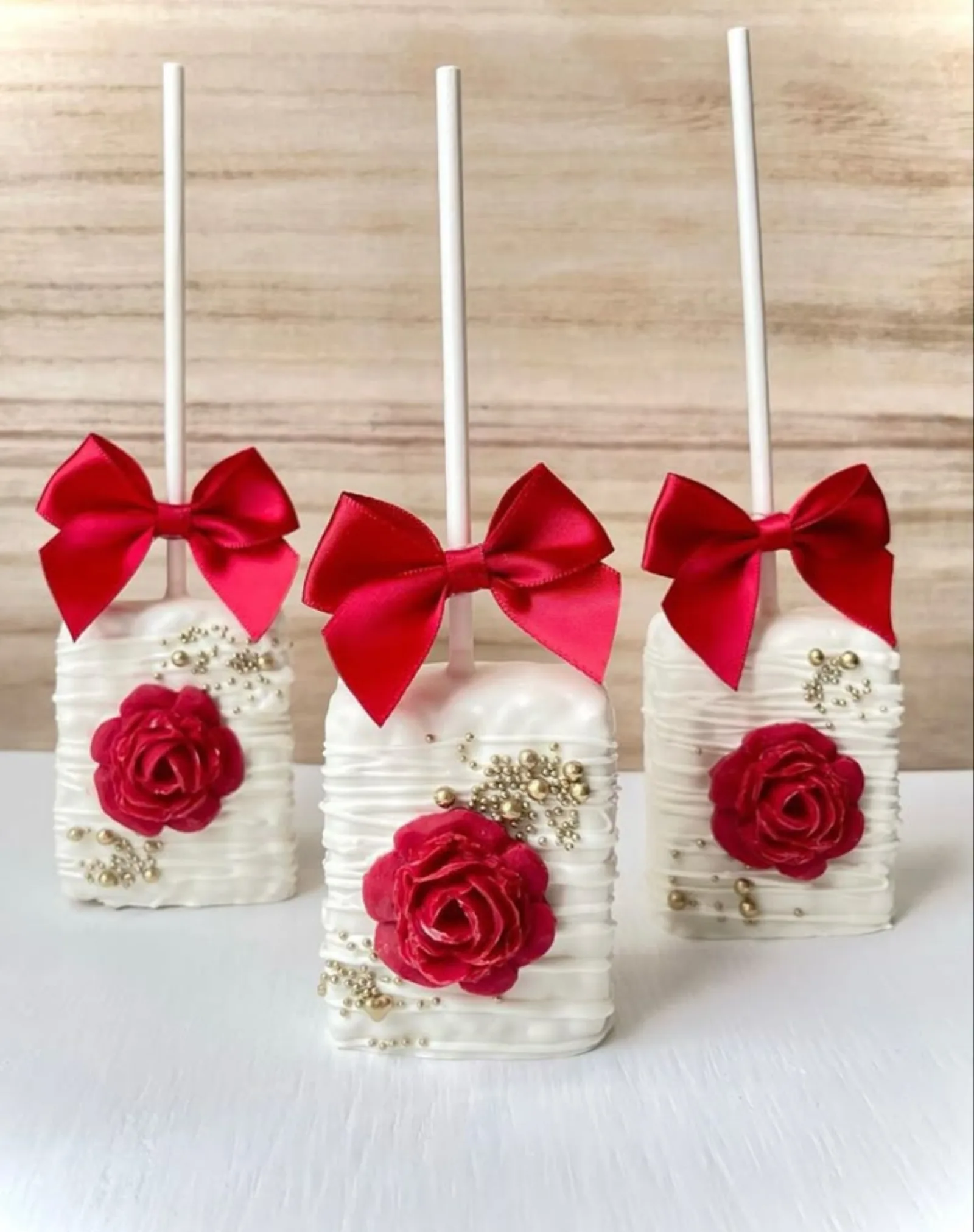 Three white chocolate cake pops with red satin bows and red fondant roses with gold pearl accents.