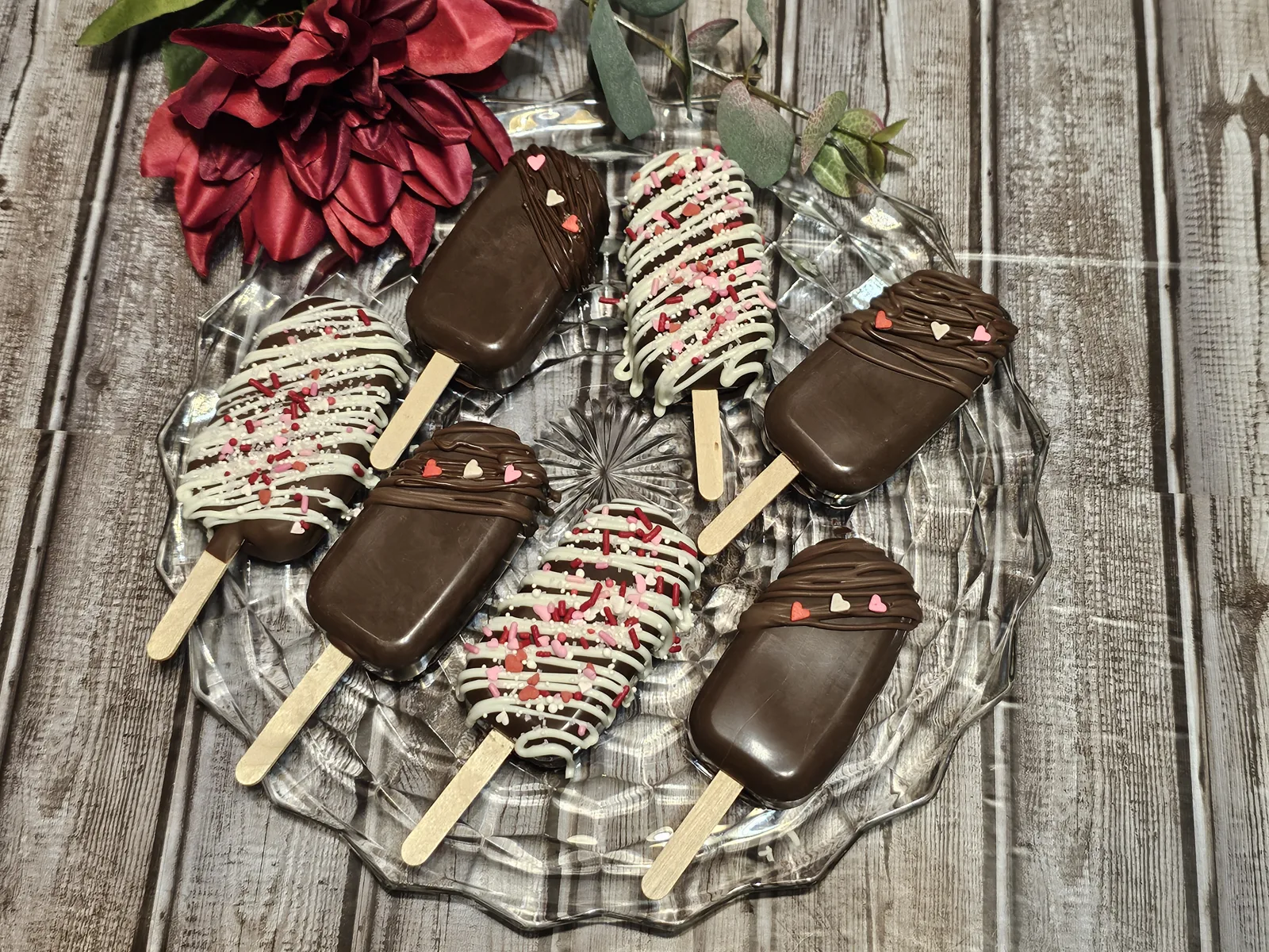 Assorted chocolate coated cakesicles on wooden sticks displayed on a glass plate, topped with white drizzle and red, pink, and white sprinkles.