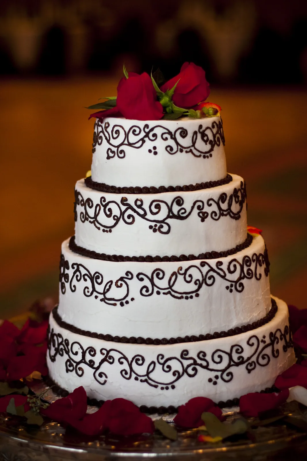 Four tier white wedding cake adorned with dark chocolate piped scrollwork, beaded borders, and topped with red roses and scattered petals.