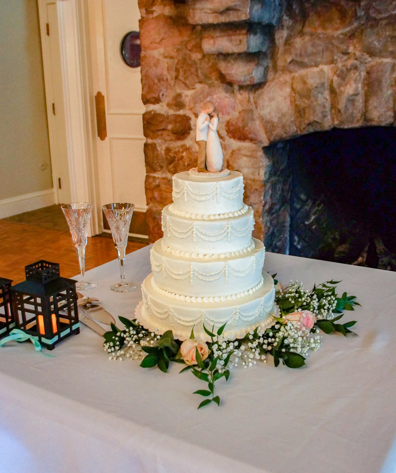 Four tiered white buttercream cake featuring elegant piped swag and pearl border designs, topped with a simple embracing couple figurine and surrounded by baby's breath and pink roses.