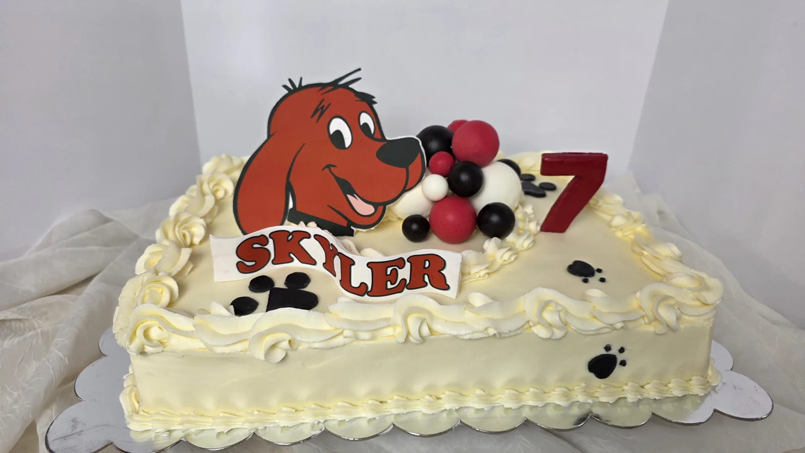 White frosted rectangular birthday cake decorated with a red dog character topper, red and black fondant balls, black paw prints, and a red number seven.