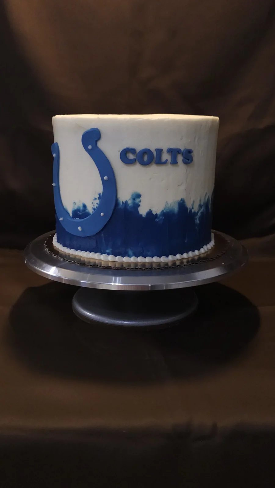 Round white frosted cake with a blue painted watercolor base, topped with a blue horseshoe emblem and COLTS lettering in royal blue.