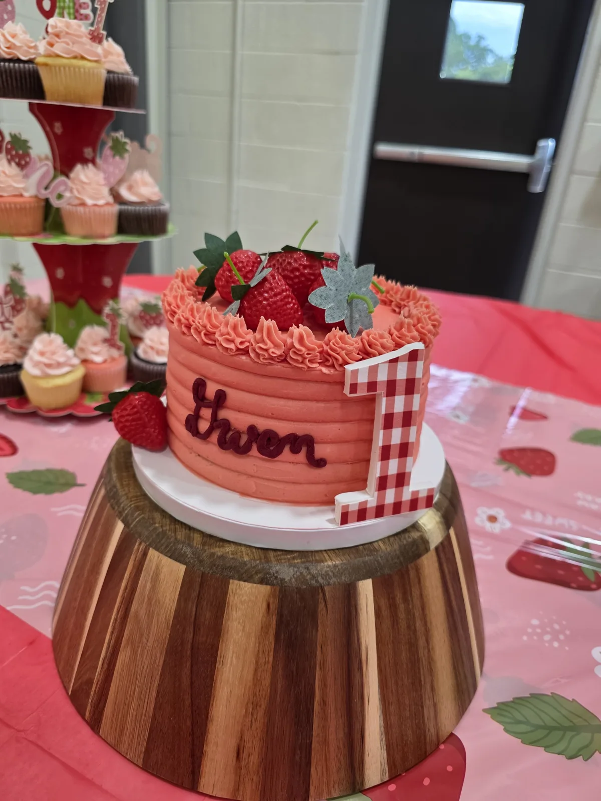 Round coral pink textured cake with ruffled frosting edge topped with fresh strawberries, silver leaves, and a red gingham number one on a wooden stand.