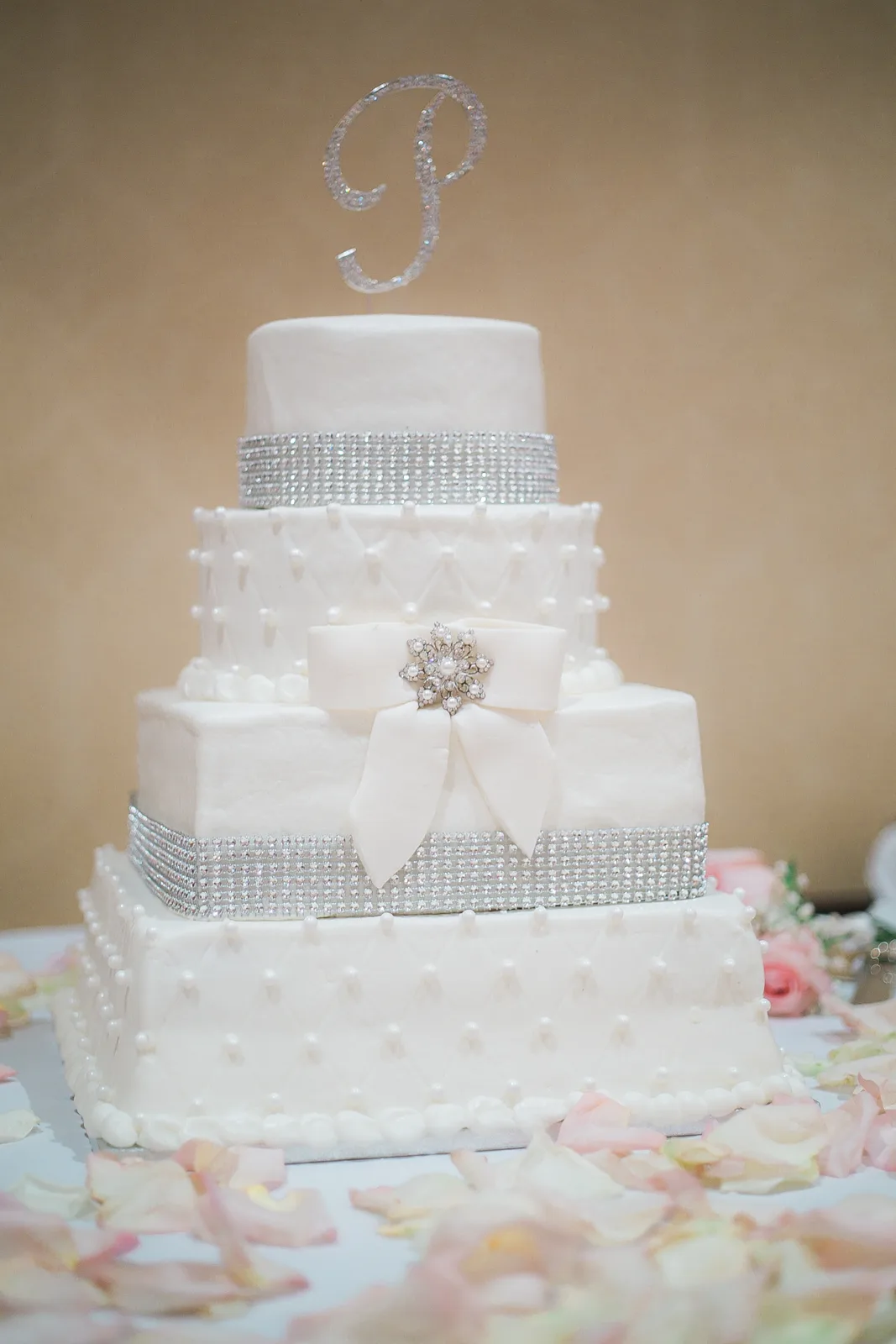 Four tier white fondant wedding cake with alternating round and square layers, silver rhinestone bands, pearl swag details, and a white bow with crystal brooch.