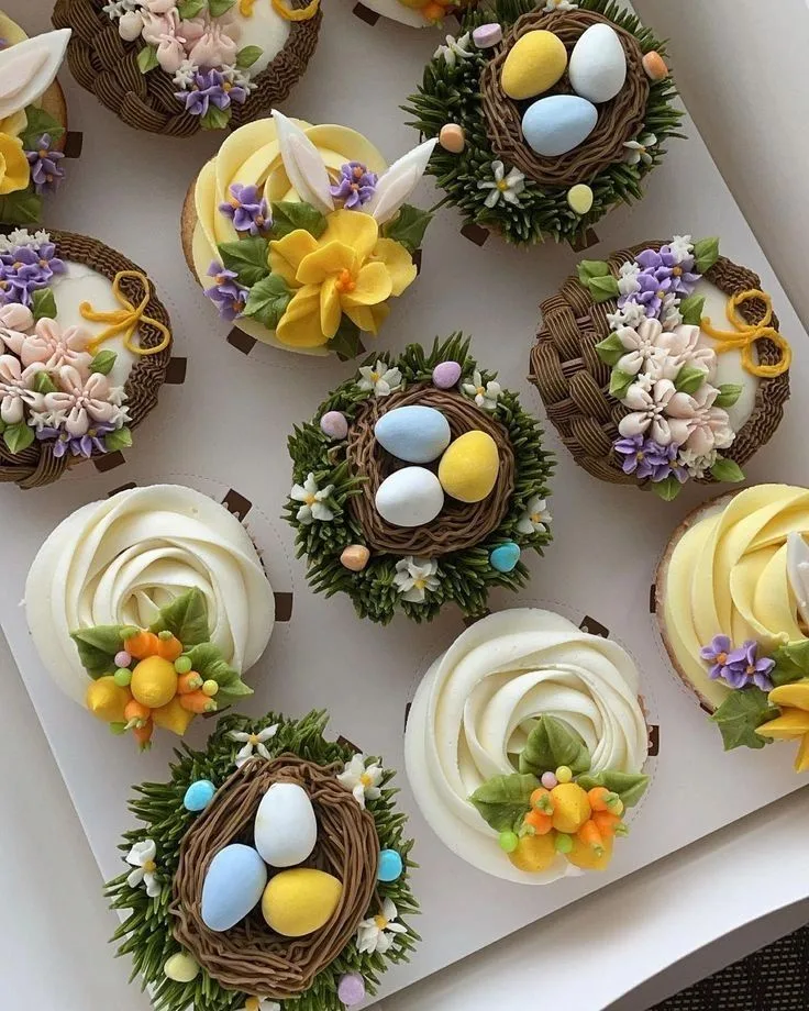 Assorted cupcakes featuring frosting nests with pastel candy eggs, yellow flowers, bunny ears, and white buttercream swirls.