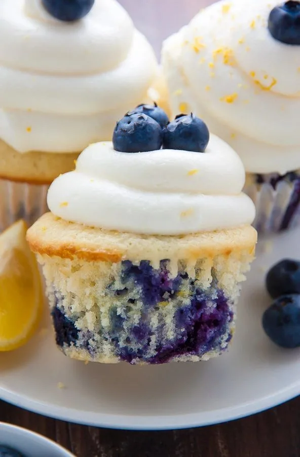 Blueberry cupcake topped with swirled white frosting and fresh blueberries, with a lemon wedge and extra berries on a white plate.
