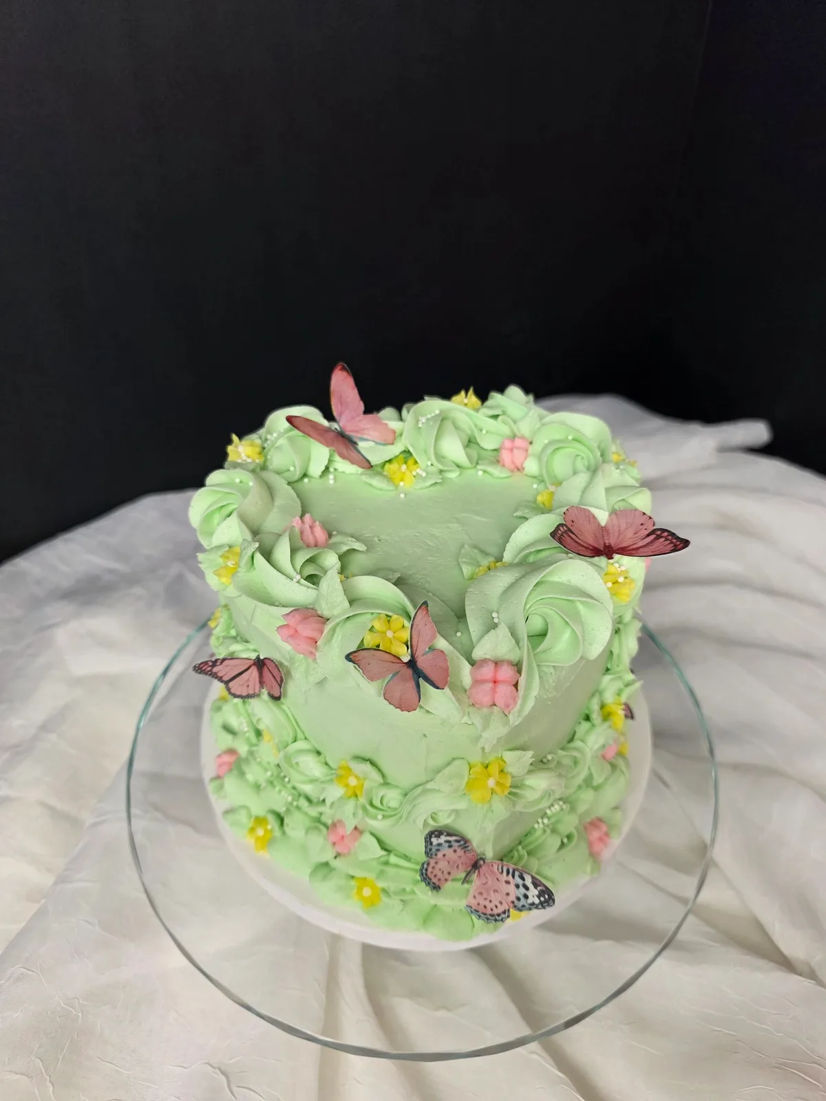 Small green frosted cake decorated with pink and yellow buttercream flowers and pink butterfly toppers on a glass stand.