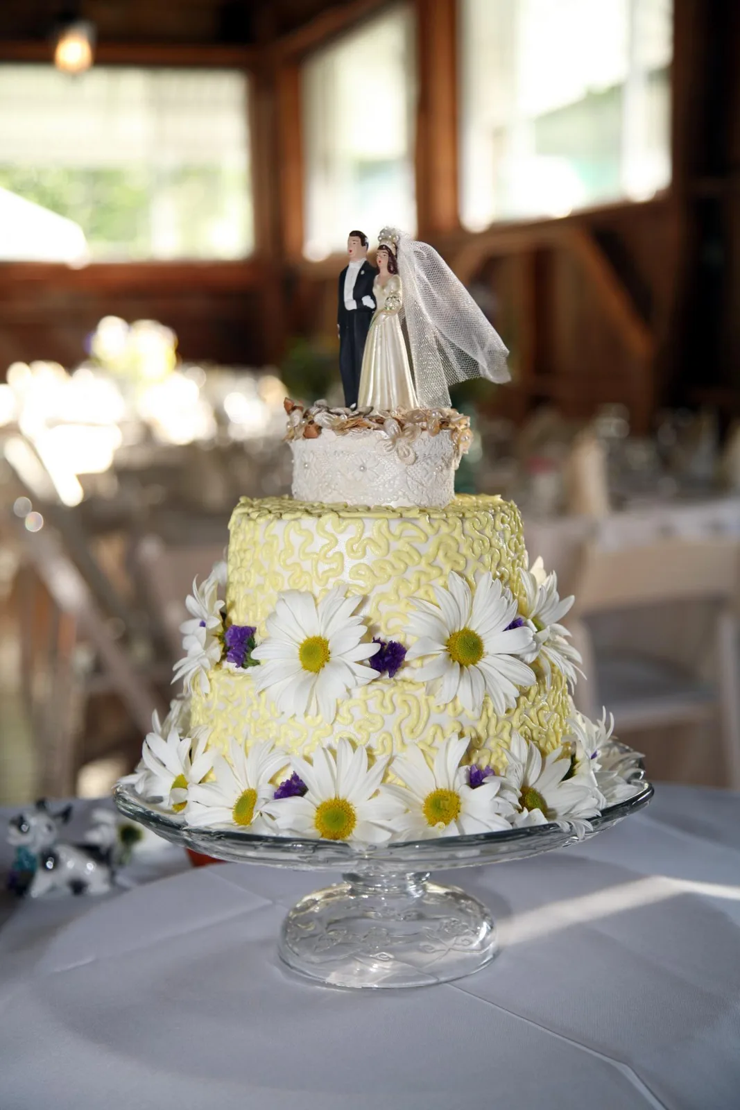 Three tiered wedding cake with white lace top tier and pale yellow scrollwork piped tiers, adorned with white daisies and purple flowers, topped with a classic bride and groom figurine.