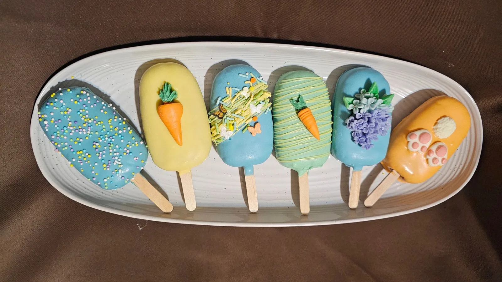 Six pastel-colored cake pops decorated with sprinkles, fondant carrots, purple flowers, and paw prints on a white oval platter.
