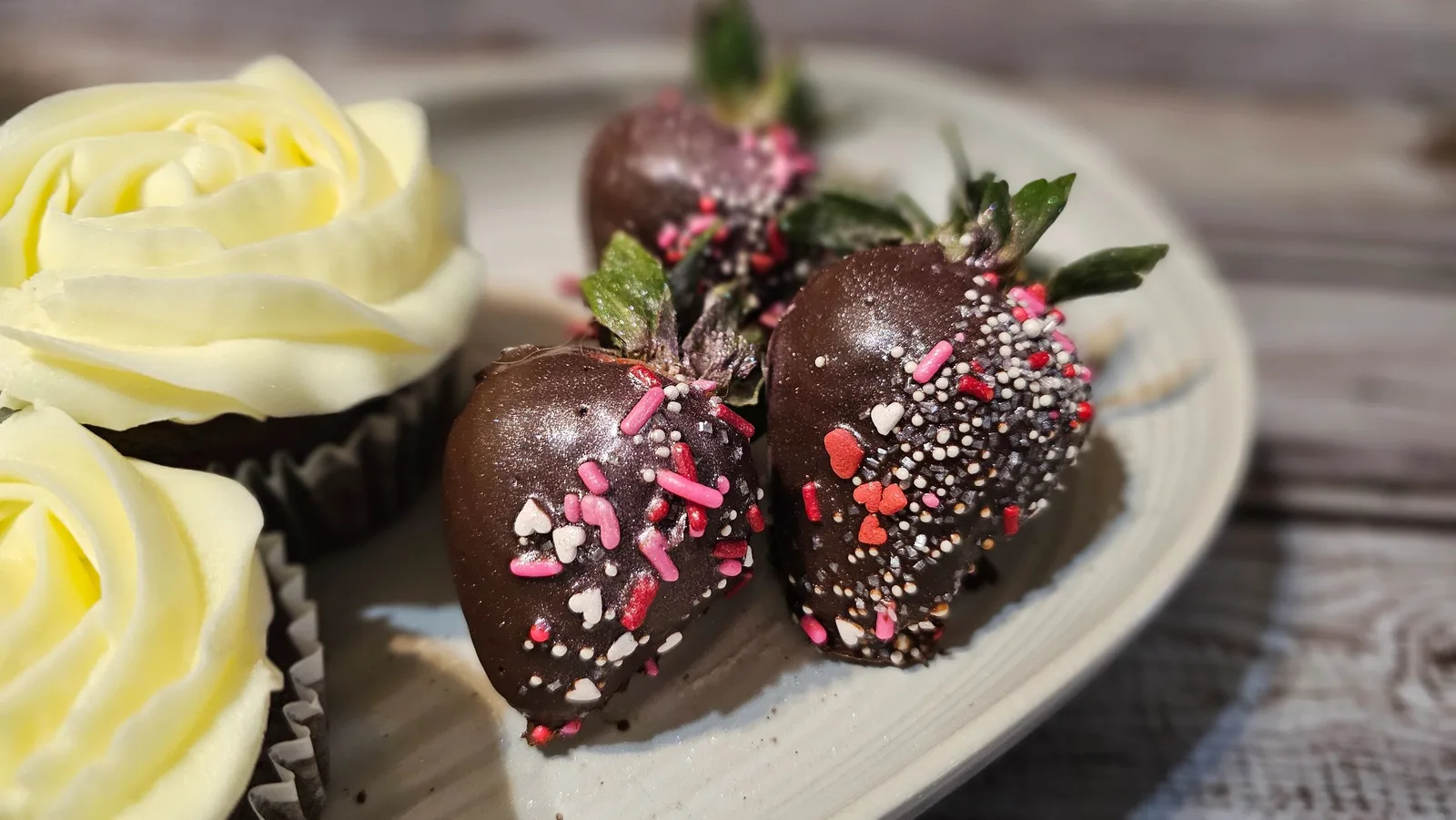 Chocolate-covered strawberries with pink, red, and white sprinkles arranged beside cupcakes topped with white buttercream roses on a plate.