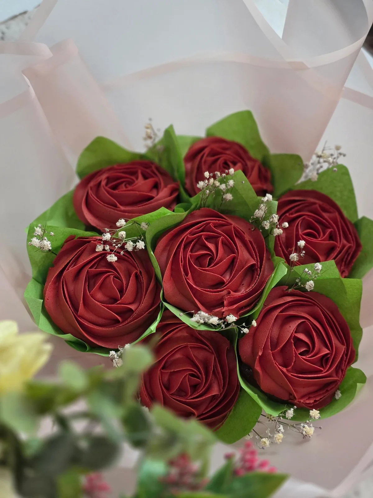 Six cupcakes topped with deep red, swirled icing shaped into roses are nestled with green paper leaves and sprigs of small white flowers.