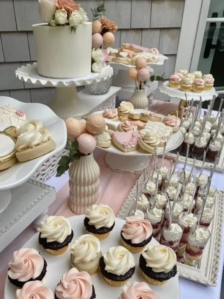 Dessert table featuring a white floral cake, frosted cupcakes, decorated cookies, cake pops, and push pops in pink and white.