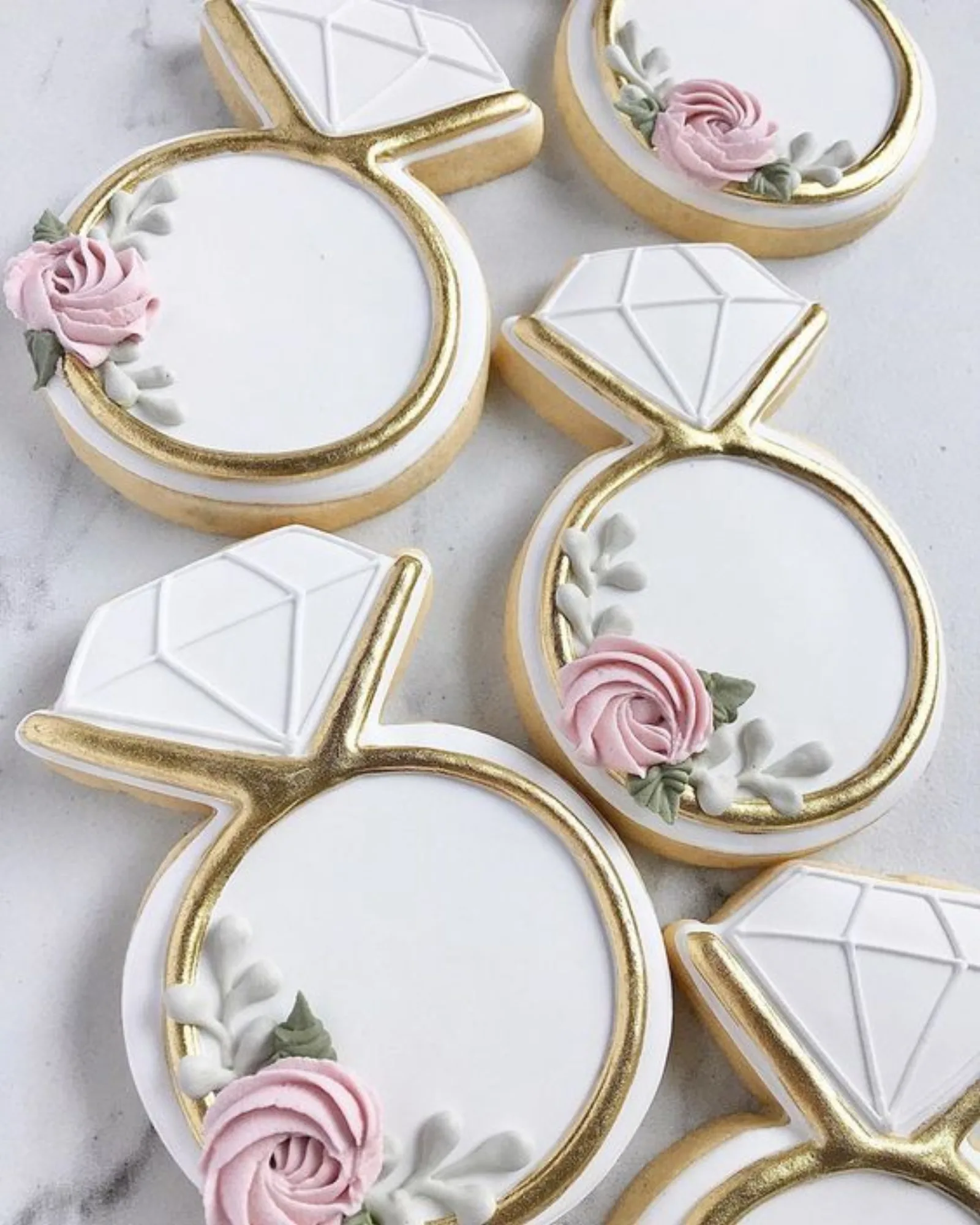 Wedding ring cookies iced in white and gold with diamond tops and piped pink roses on marble.