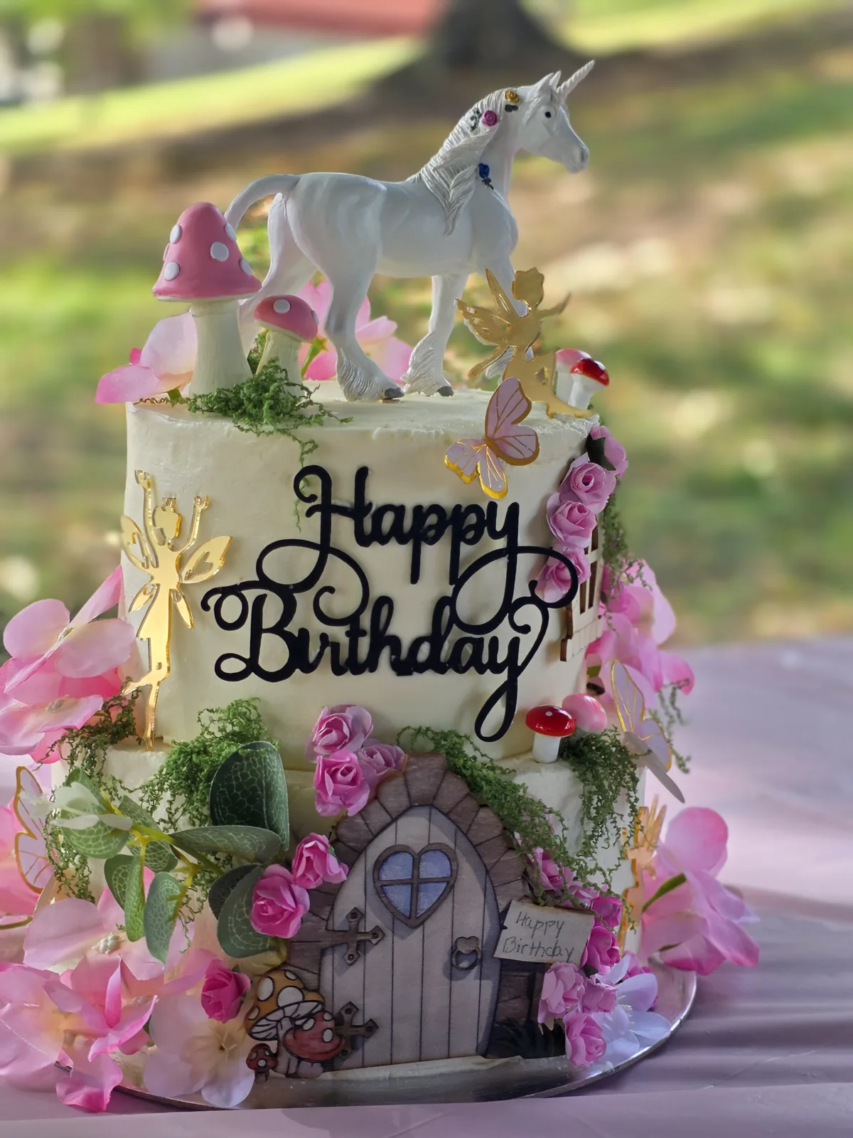 Two-tier fairy garden birthday cake with a unicorn topper, pink roses, toadstools, gold fairies and a painted fairy door.